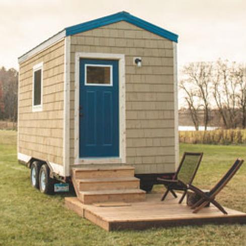 A tiny house parked in a field with a small deck and two chairs in front.